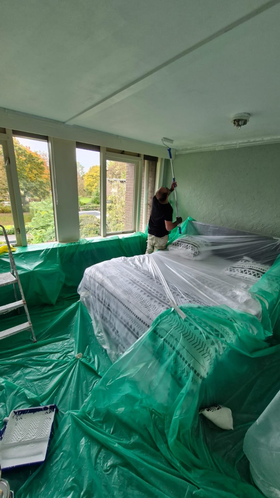 Slaapkamer plafond en wanden schilderen | Nour Klusbedrijf Schilder verft het slaapkamerplafond met een verfroller boven afgedekte meubels